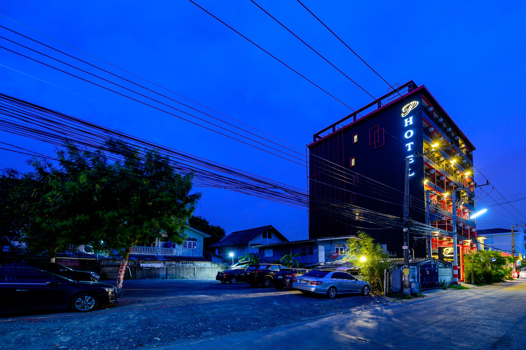 Un hotel de varios pisos con un letrero llamativo, coches aparcados y un árbol en primer plano.