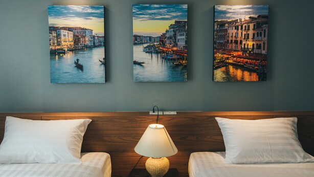 Une chambre d’hôtel avec un lit, deux oreillers, une lampe et trois tableaux représentant un paysage urbain accrochés au mur.