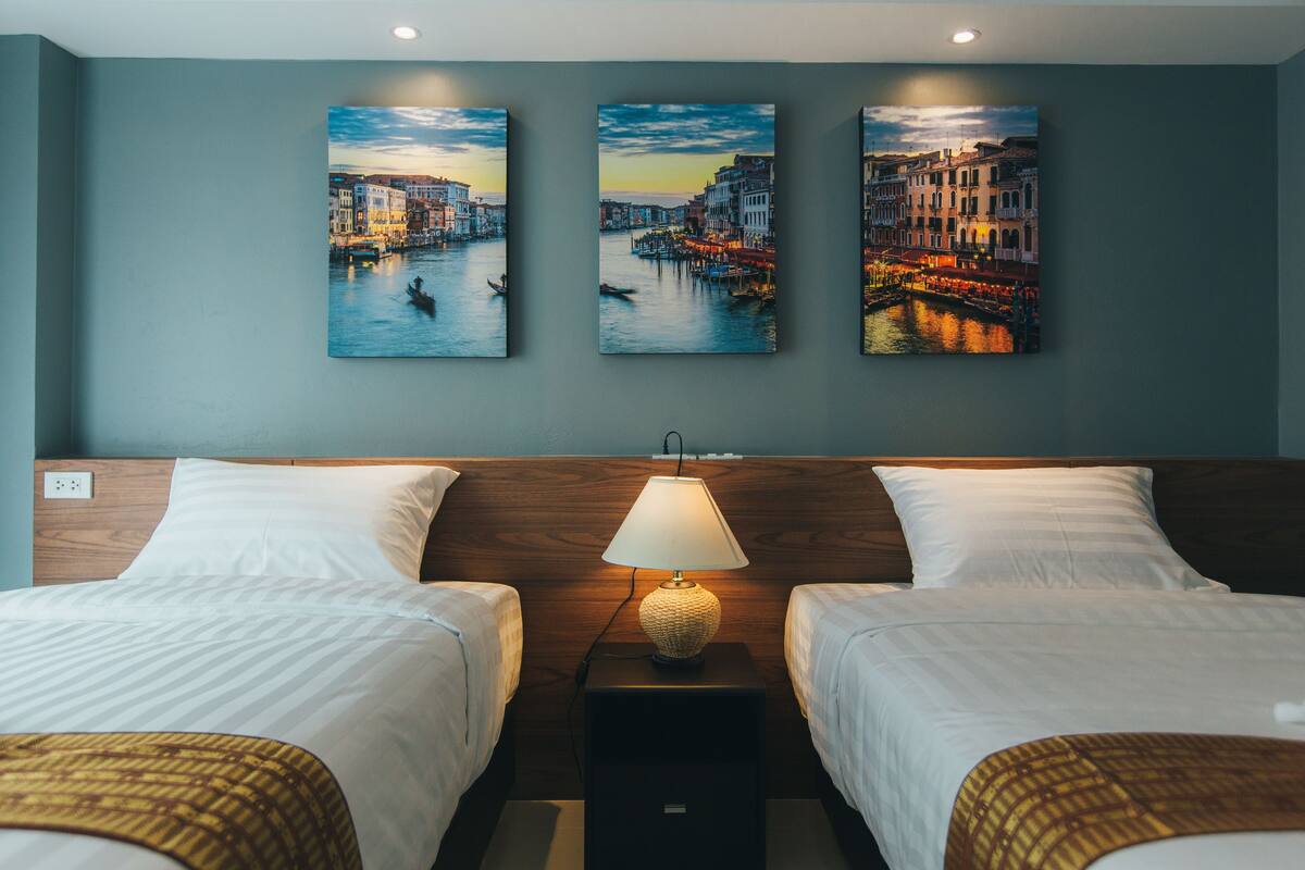 A hotel room with two beds, a nightstand with a lamp, and three paintings of a cityscape on the wall.