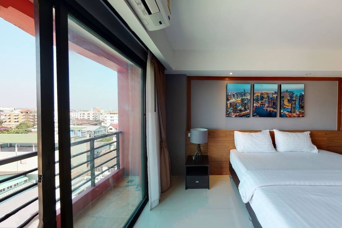 A hotel room with a large bed, a nightstand, a lamp, and a cityscape view through the window.