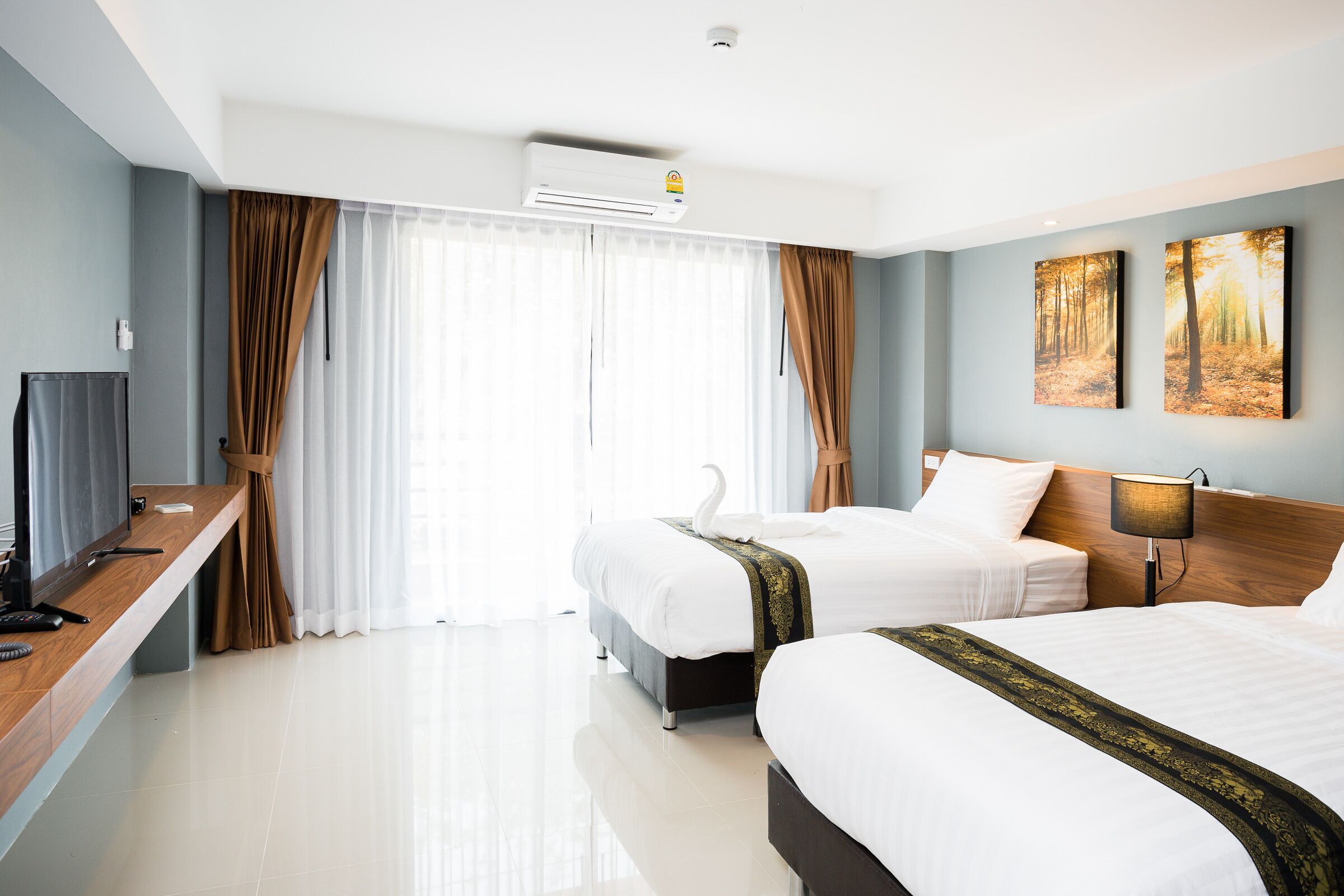 A hotel room with two beds, a desk with a TV, and a large window with curtains.