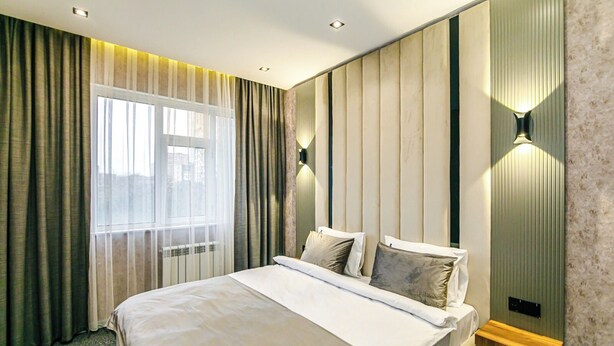 Una habitación de hotel moderna con una cama grande, una ventana con cortinas y una lámpara fijada a la pared.