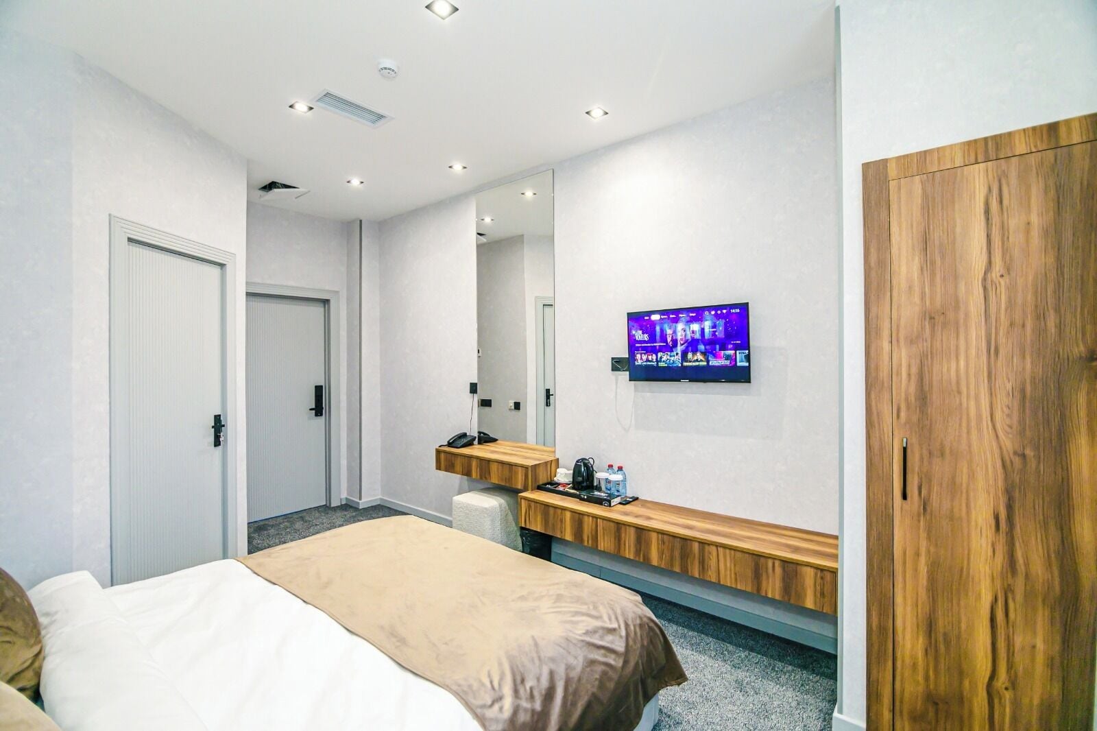 A modern hotel room with a bed, a wooden desk, a television mounted on the wall, and a wardrobe.