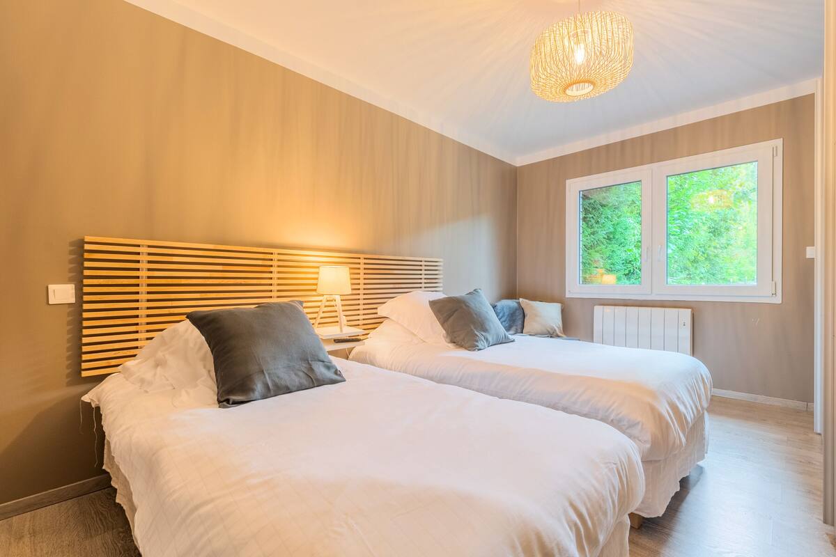 Quarto com duas camas, cabeceira de madeira, abajur e janela com vista para vegetação.