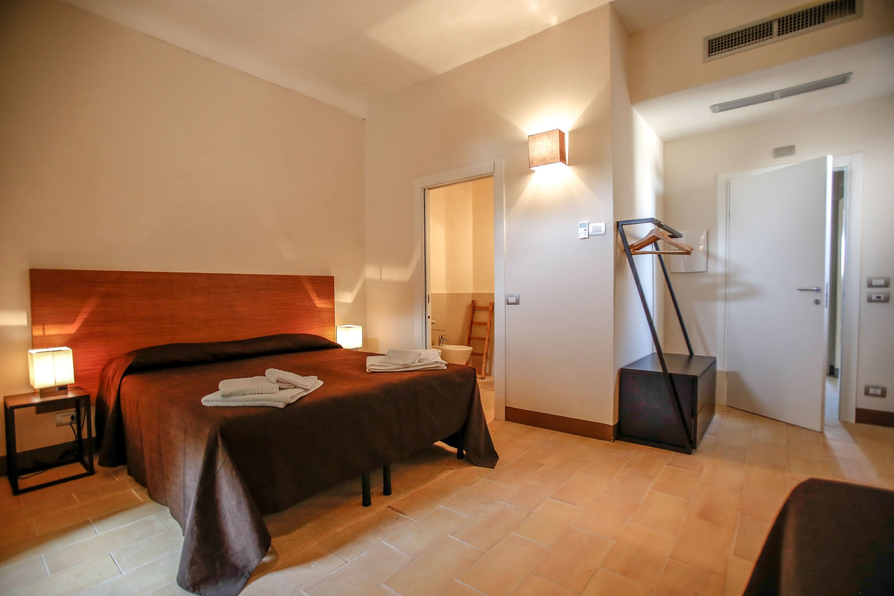 A hotel room with a large bed, bedside table with a lamp, a wardrobe, and a bathroom visible through an open door.
