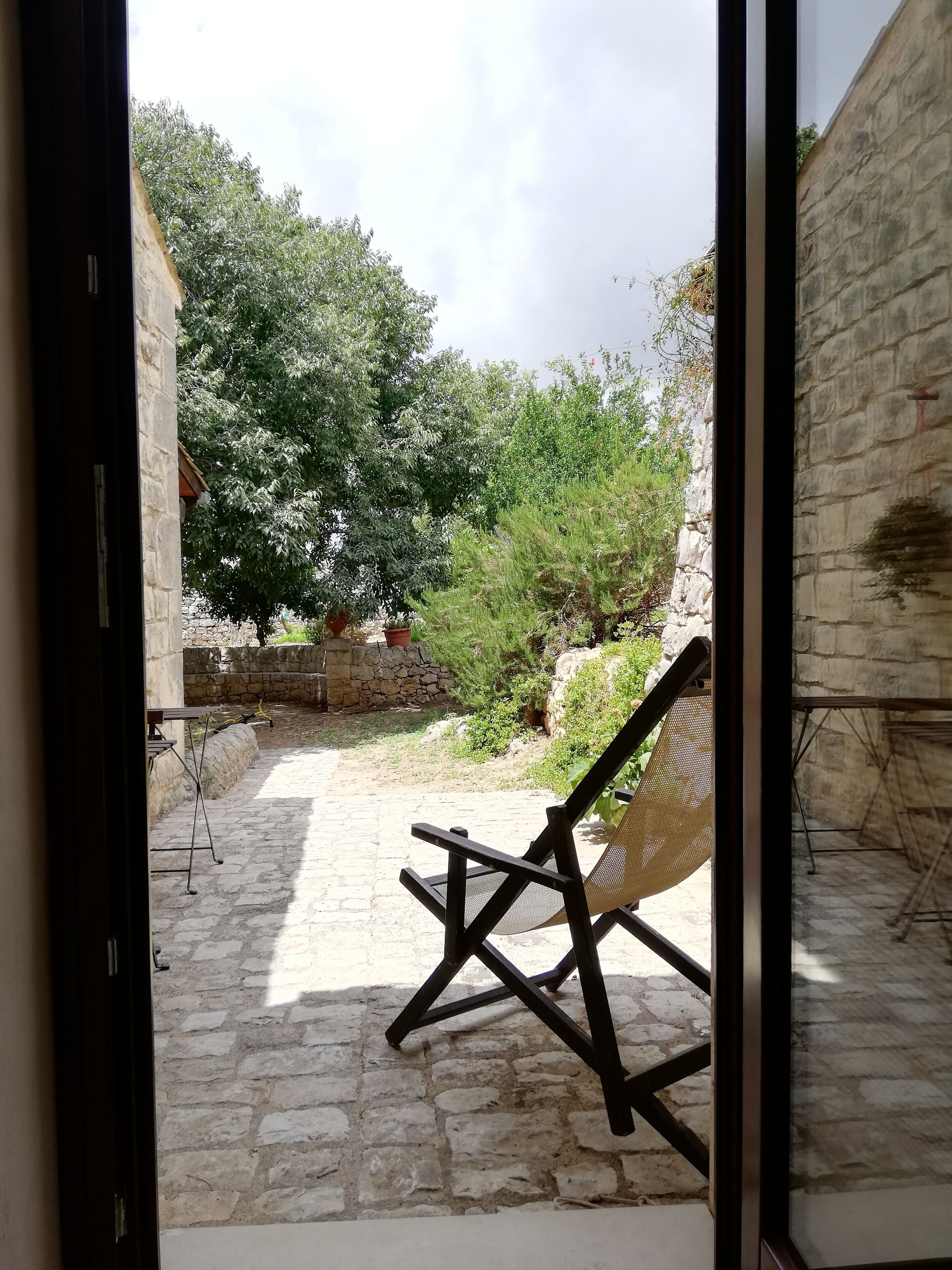 A stone patio with a wicker chair and a table, surrounded by greenery and a stone wall.