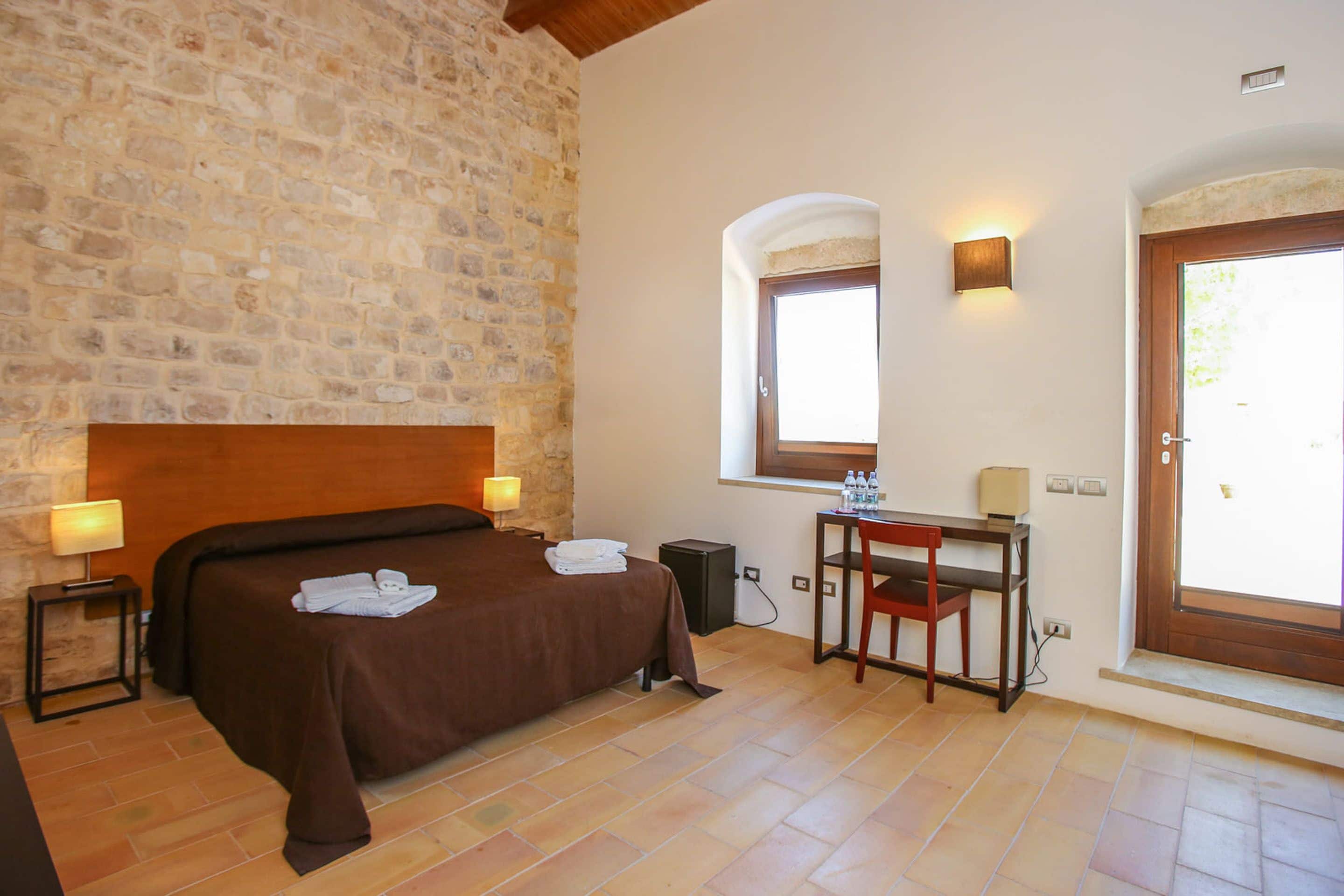 Ein Hotelzimmer mit Steinwand, einem Bett mit brauner Kopfteil, einem Schreibtisch mit Stuhl und einem Fenster mit Blick.