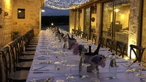 A long dining table set for a meal with string lights overhead, stone walls, and a view of the landscape. - Le Chiuse di Guadagna (Scicli)