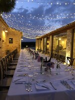 Una larga mesa de comedor dispuesta para una comida, con luces colgantes, paredes de piedra y vistas al paisaje.