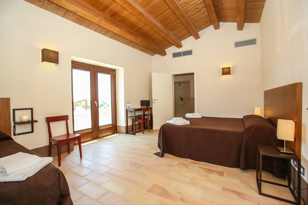 A hotel room with a bed, a desk, a chair, a shower, and a small table with a lamp.