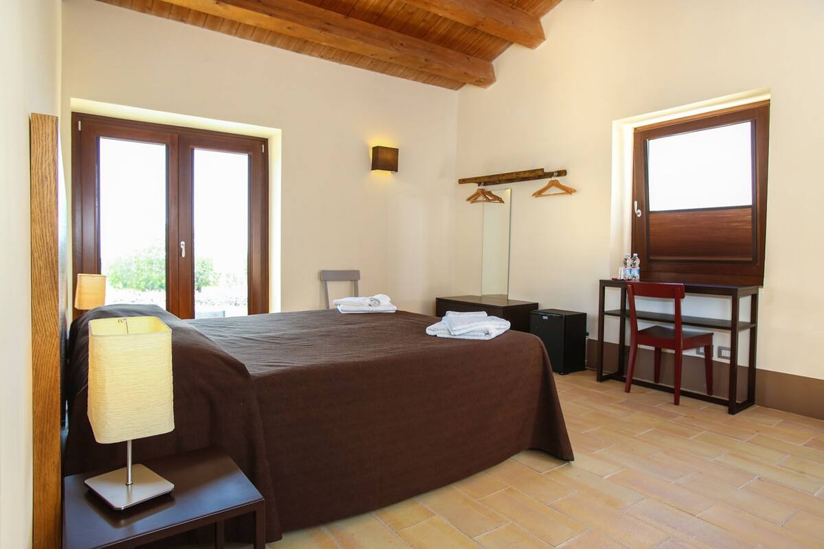 A bedroom with a bed, bedside table, lamp, chair, desk, and two windows.