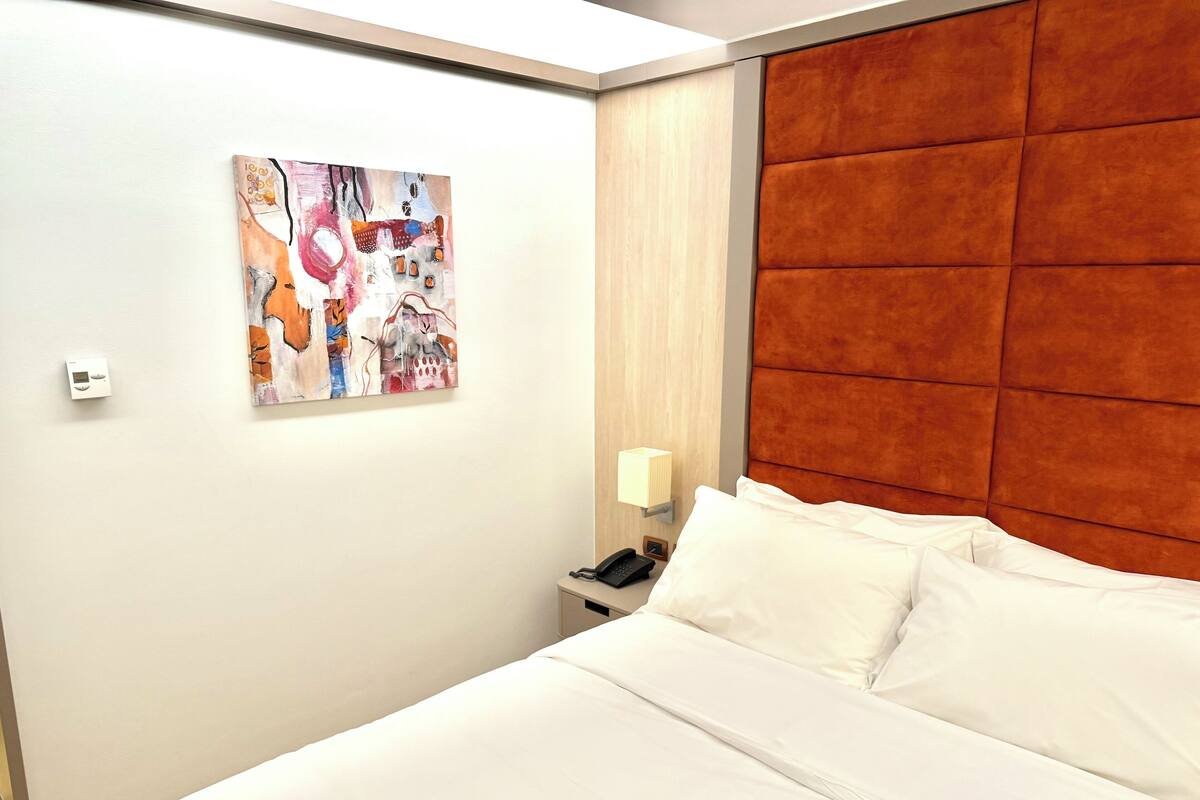 A hotel room with a large bed, a nightstand, a telephone, a wall-mounted light, and a colorful abstract painting.