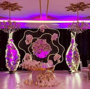 A wedding setup with a central floral arrangement, two side floral columns, and a table with glasses and a bottle.