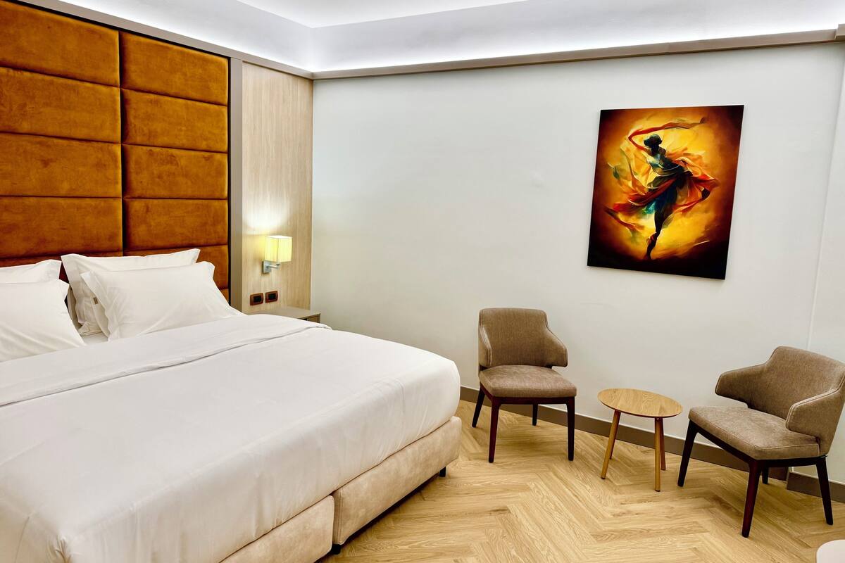 A hotel room with a large bed, two armchairs, a small table, a painting on the wall, and a nightstand with a lamp.