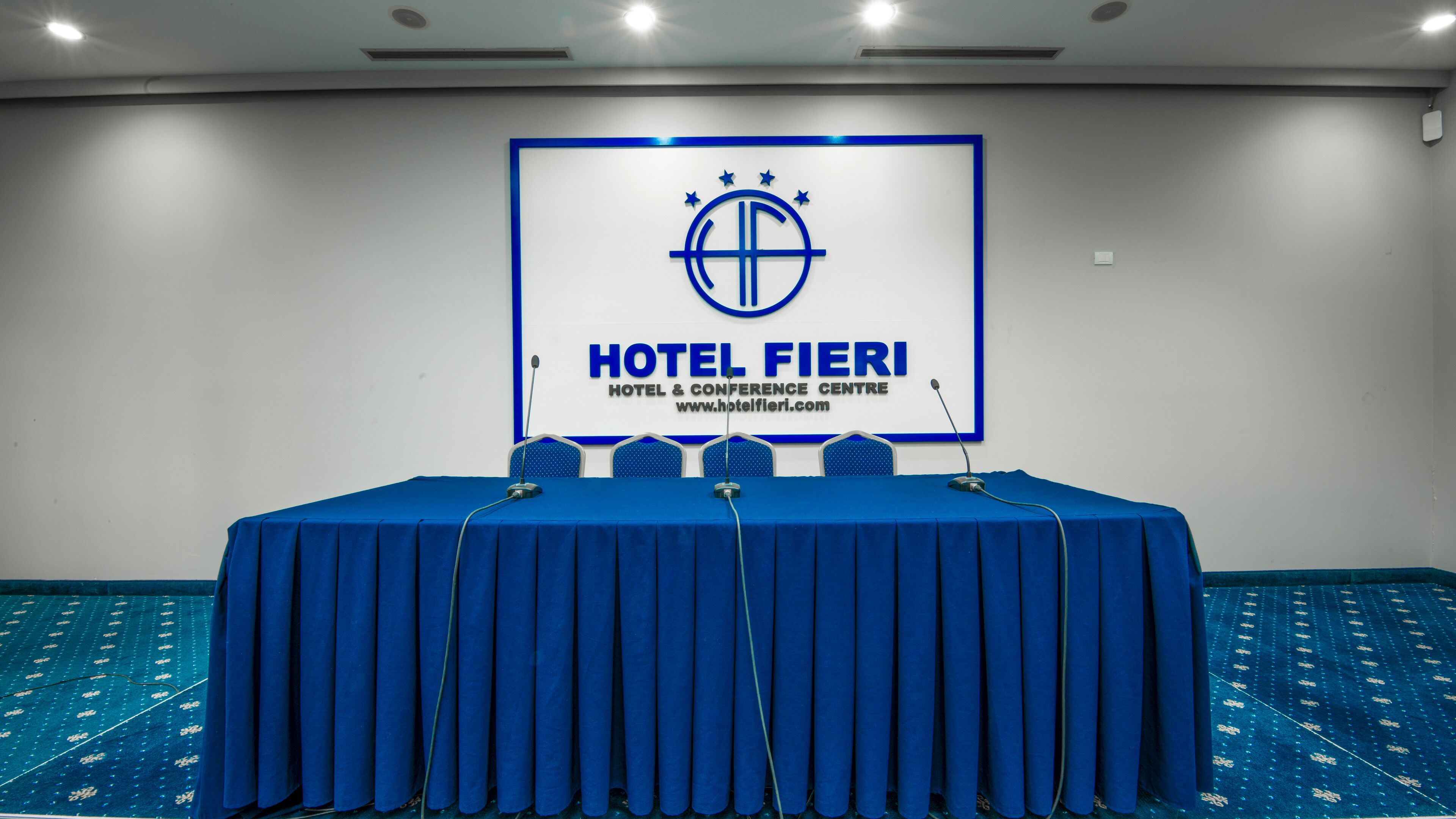 Una sala de conferencias en el Hotel Fieri, con una mesa y sillas azules, un atril y un logotipo en la pared.