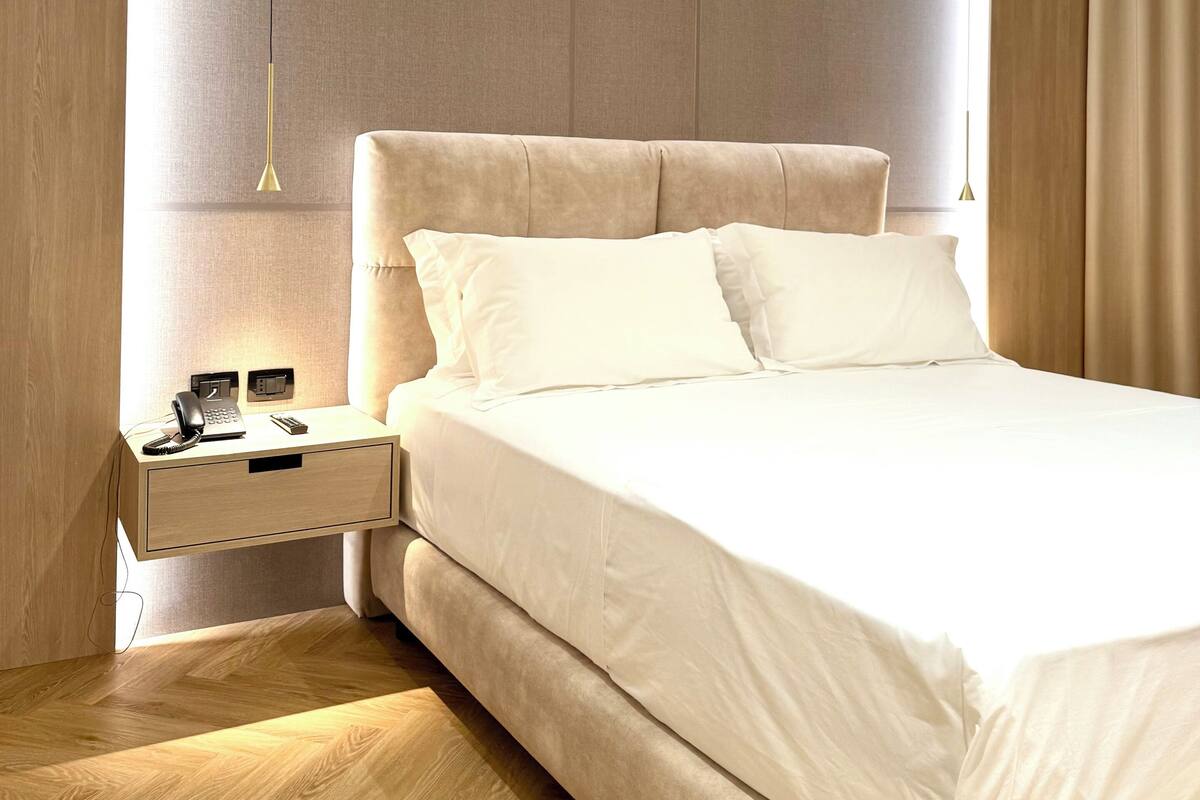 A modern bedroom with a large bed, a bedside table with a phone and a lamp, and a wooden floor.