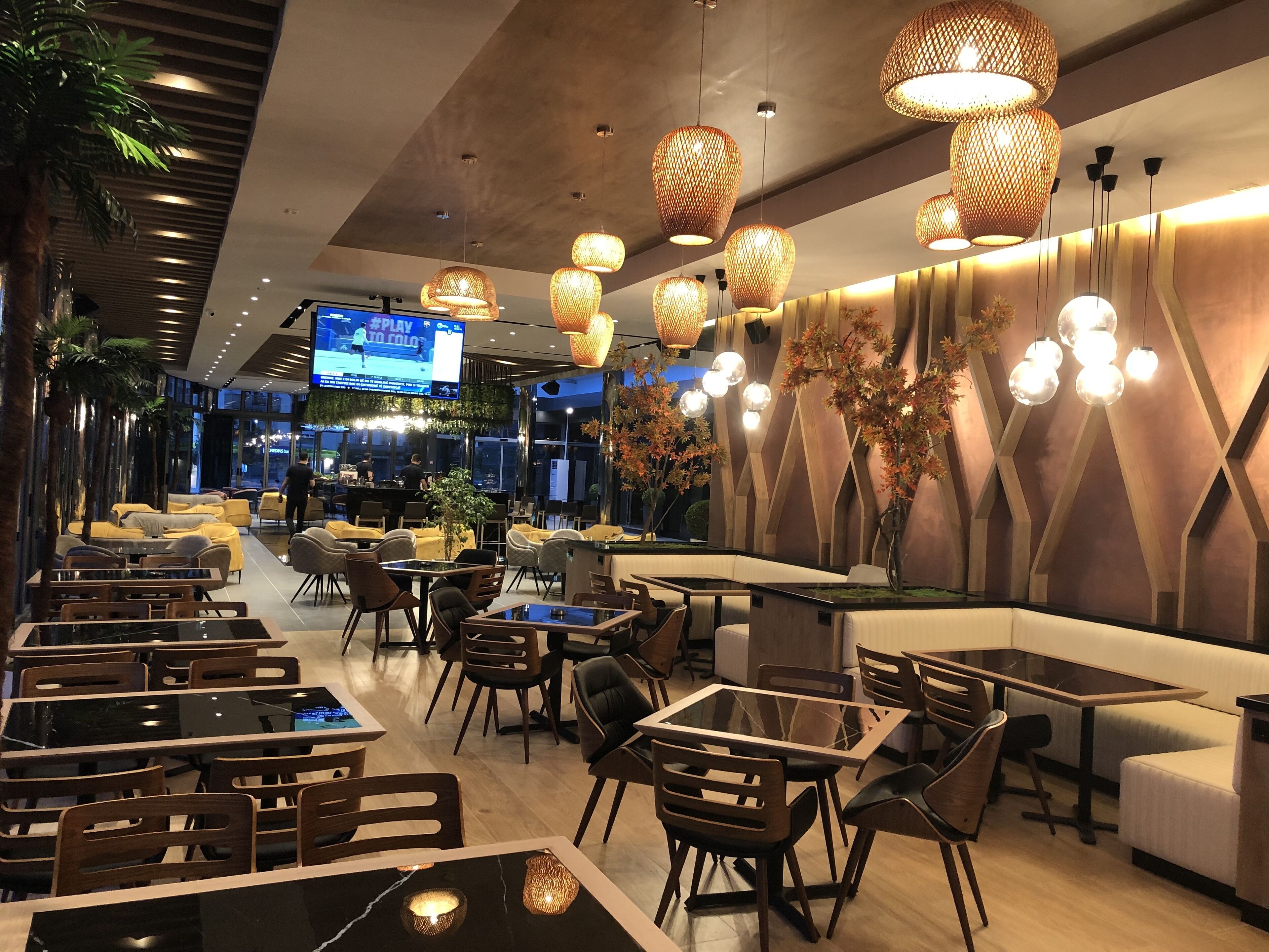 Un restaurante moderno con diversas opciones de asientos, incluyendo mesas, sillas y cabinas. Hay luces decorativas colgantes y una pantalla grande que muestra un evento deportivo.
