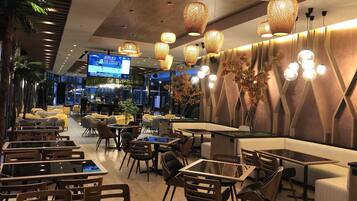 Un restaurante moderno con diversas opciones de asientos, incluyendo mesas, sillas y cabinas. Hay luces decorativas colgantes y una pantalla grande que muestra un evento deportivo.