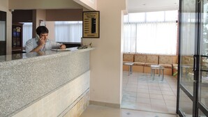 A hotel reception area with a receptionist, a framed room rate sign, and a seating area with a view of the outdoors. - Acikgoz Hotel (Edirne)