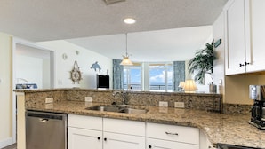 A kitchen with granite countertops, white cabinets, a stainless steel dishwasher, and a view of the ocean through large windows.