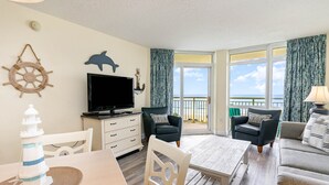 A living room with a TV, a dining table, and a view of the ocean through large windows.