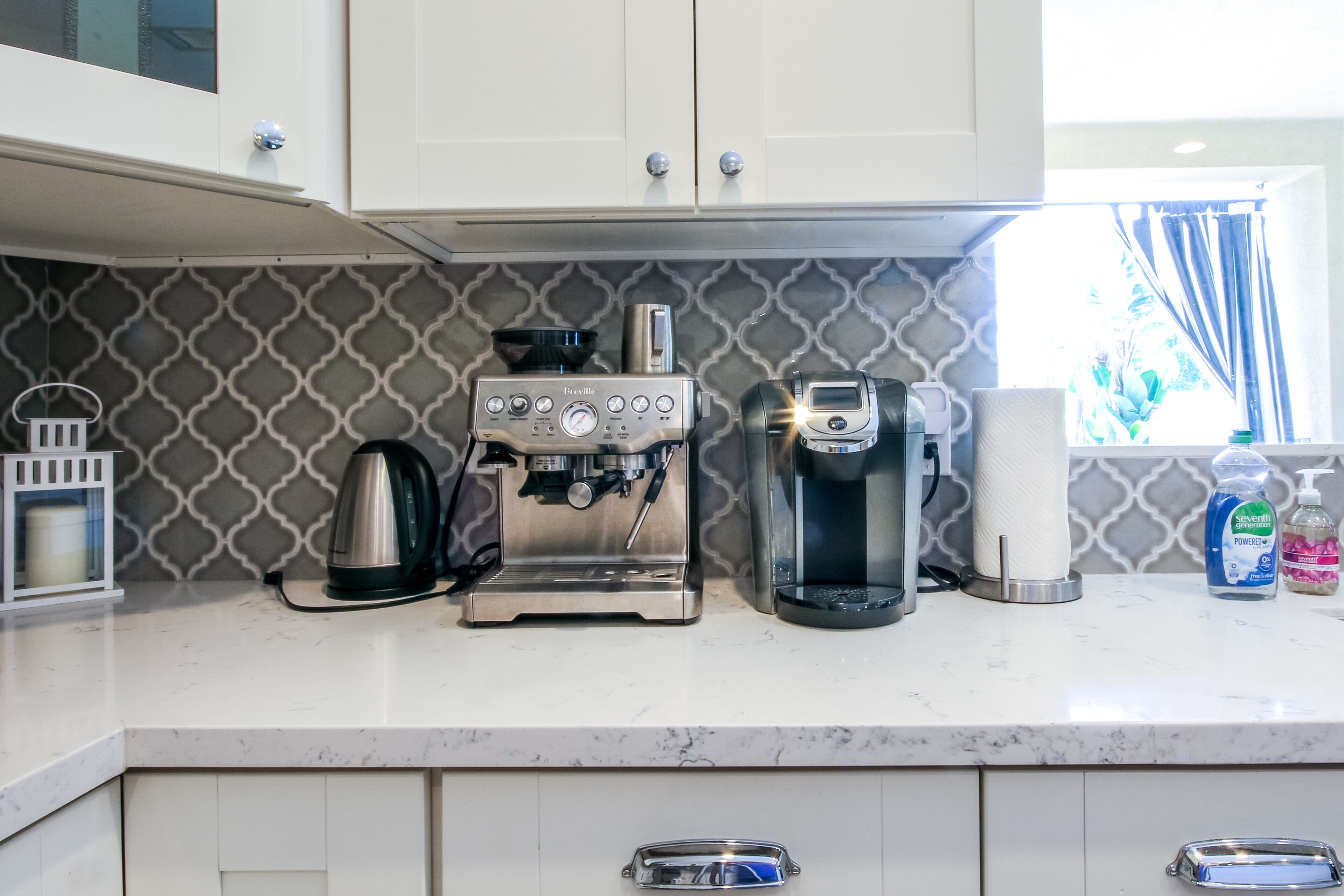 Een keuken met een koffieapparaat, waterkoker en koffiemachine. Er is een raam met gordijnen, een handzeepfles en een papieren handdoekhouder.