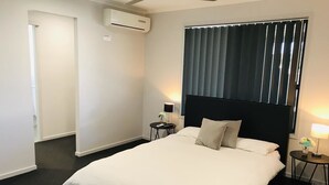A hotel room with a large bed, a ceiling fan, an air conditioning unit, a window with blinds, and two bedside tables with lamps. - Hervey Bay Holiday Home (Kawungan)