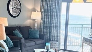 A living room with a sofa, coffee table, and dining table set with green bowls and plates. A large window offers a view of the ocean. - 3Bd Condo in Beachside Resort w/ Amenities Galore! (Myrtle Beach)