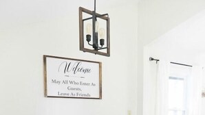 Une table de salle à manger en bois, accompagnée d’un banc, d’une pancarte d’accueil encadrée et d’un luminaire suspendu.