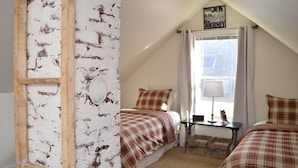 Une chambre confortable avec un plafond en pente, une échelle en bois, deux lits avec une literie à carreaux, une petite table avec une lampe et une fenêtre avec des rideaux.