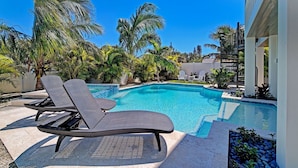 A swimming pool with lounge chairs, surrounded by palm trees and a building.