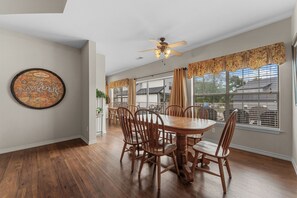 Un coin repas avec une table et des chaises en bois, un ventilateur de plafond et une grande fenêtre avec des rideaux.