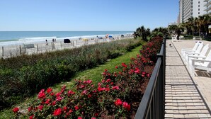 A coastal promenade with a beach, a row of white chairs, and a tall hotel building.