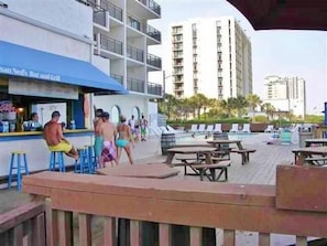 Ein Poolbereich mit Bar und Restaurant, Sitzgelegenheiten im Freien und Blick auf ein hohes Gebäude im Hintergrund.