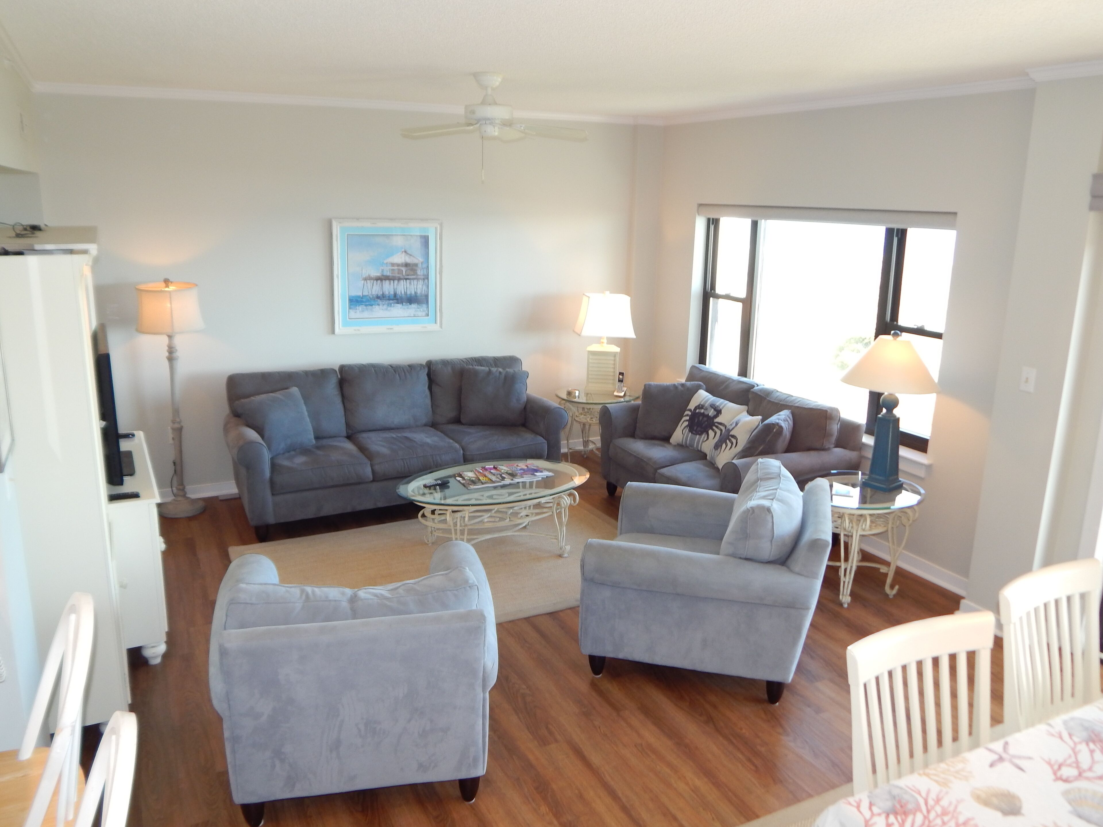 A living room with a grey sofa, two armchairs, a coffee table, and a ceiling fan. There is a window with blinds and a painting on the wall.