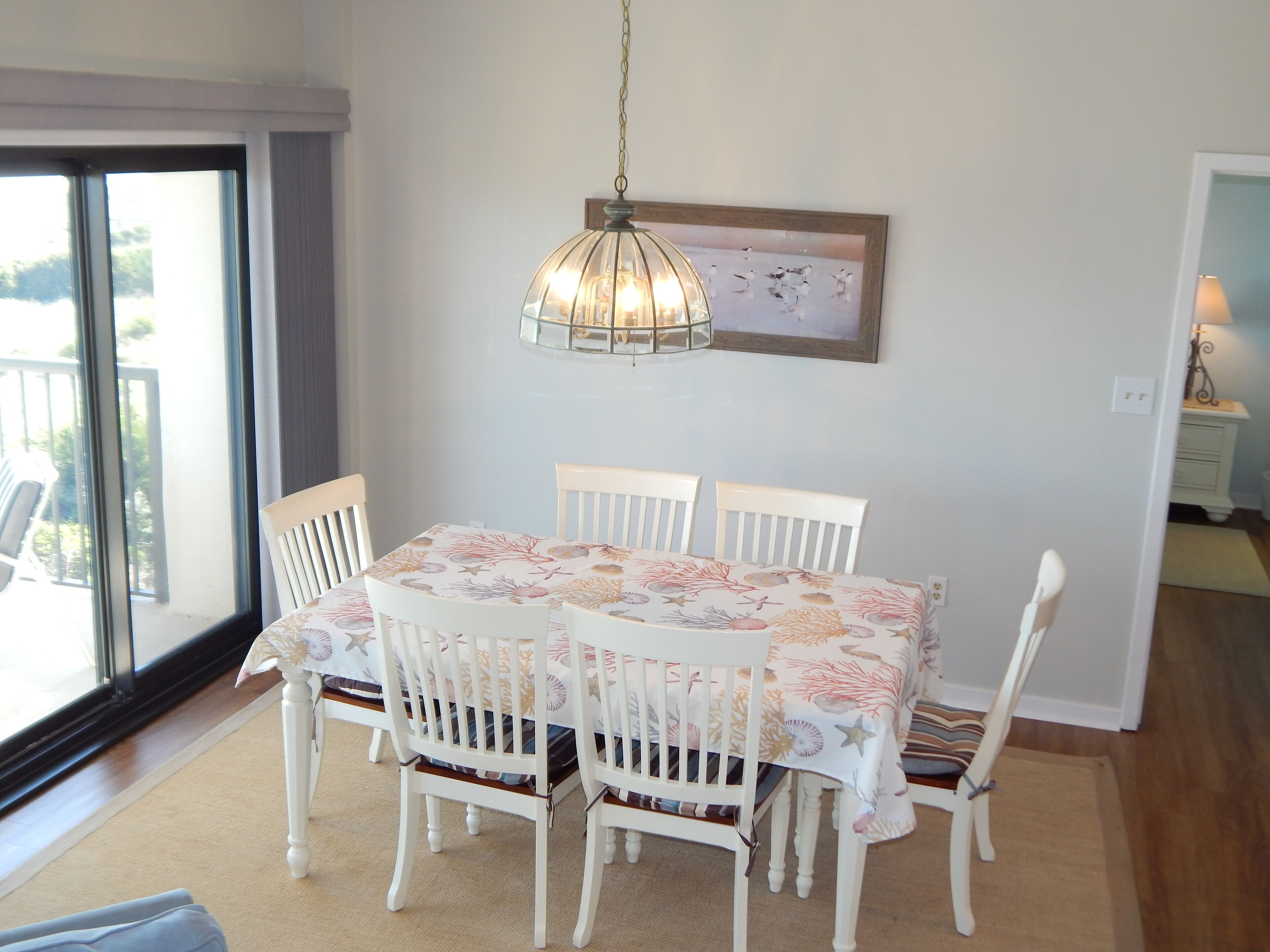 A dining area with a table set for four, white chairs, a hanging light fixture, and a framed picture on the wall.