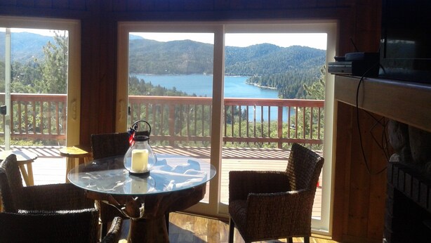 Une pièce avec une table en verre et des chaises en osier, une bougie, et une vue sur un lac et des montagnes, visible à travers de grandes fenêtres.
