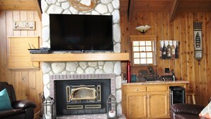 L’intérieur chaleureux d’un chalet avec une cheminée en pierre, un téléviseur à écran plat fixé au-dessus, des murs en bois et un coin cuisine doté d’un plan de travail en pierre.