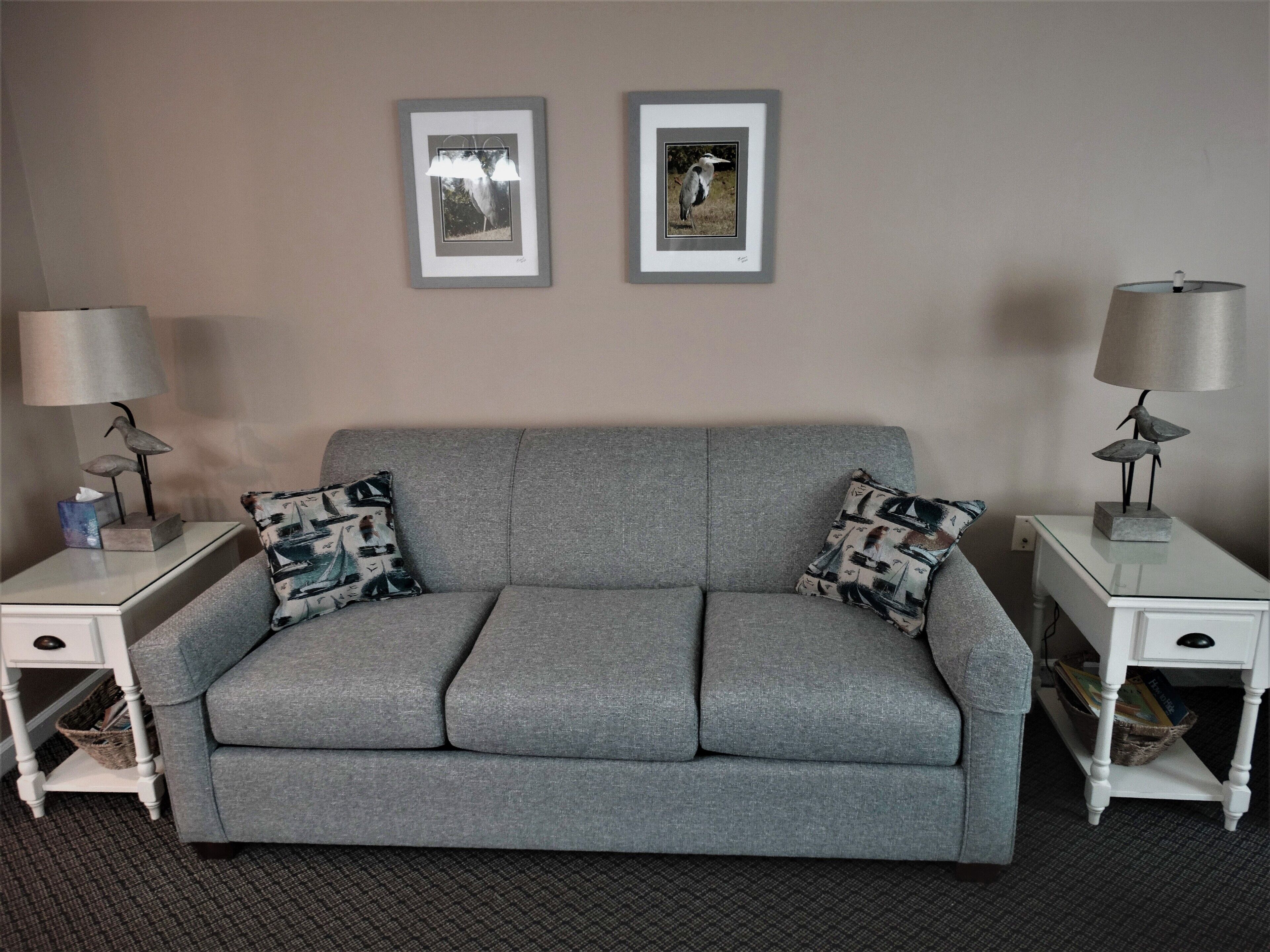 A grey sofa with two patterned pillows, flanked by white nightstands with lamps and a basket. Two framed pictures hang on the wall above.