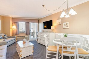 Een hotelkamer met een bank, een salontafel, een eettafel met stoelen, een televisie, een lamp en een raam met gordijnen.
