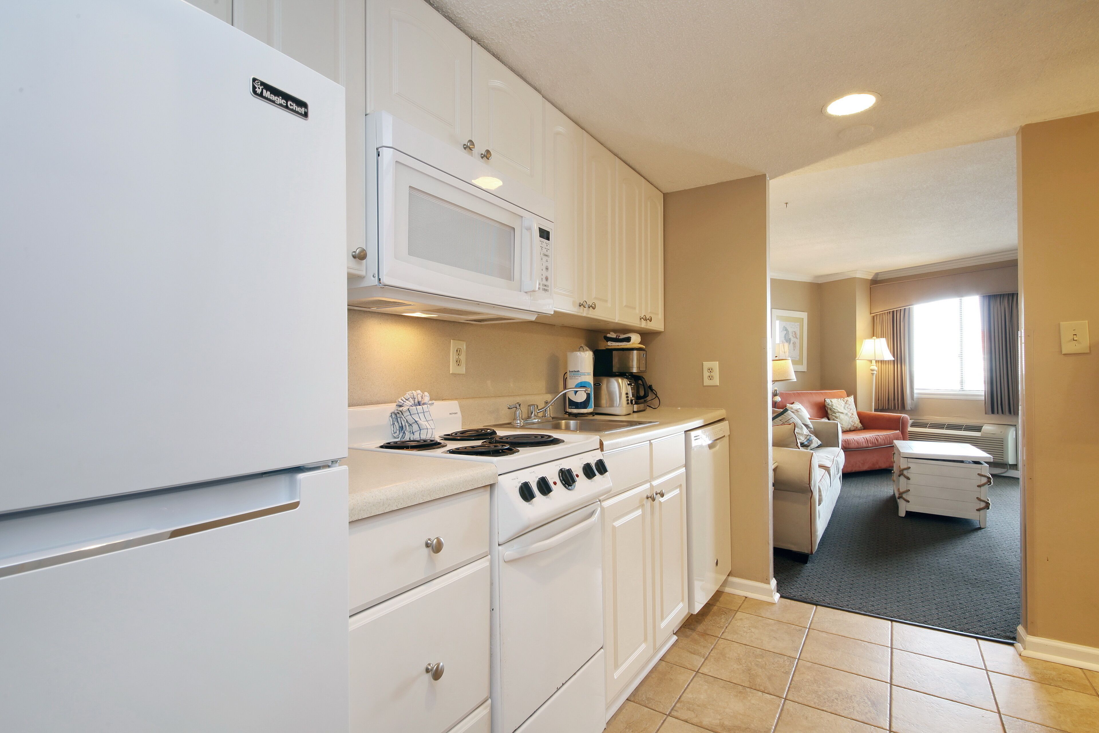 A compact kitchen with white appliances, a microwave, stove, and a coffee maker. There is a small dining area with a table and chairs, and a living room with a sofa and a lamp.