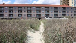 A multi-story hotel with a red roof and balconies, surrounded by tall grass and a sandy path.