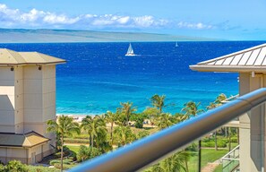 A coastal view with a sailboat on the water, palm trees, and a building with a balcony.