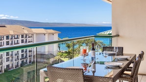 A balcony with a glass table set for dining, overlooking a sea view with buildings in the background.