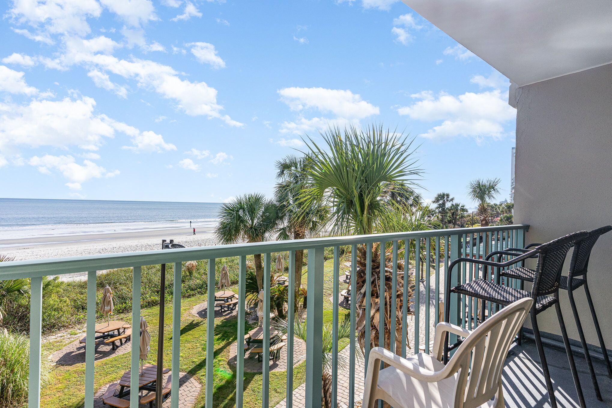 A balcony with a view of the beach, outdoor seating, and palm trees.