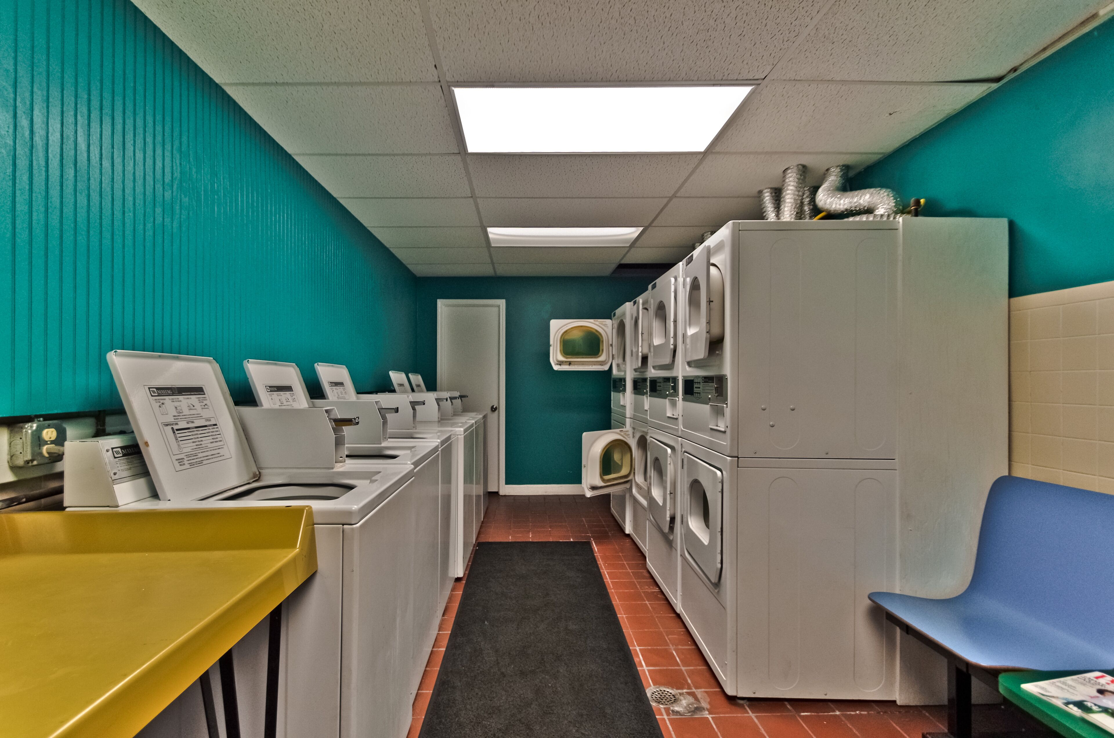 A laundry room with multiple washing machines and dryers, a yellow sink, and a blue bench.