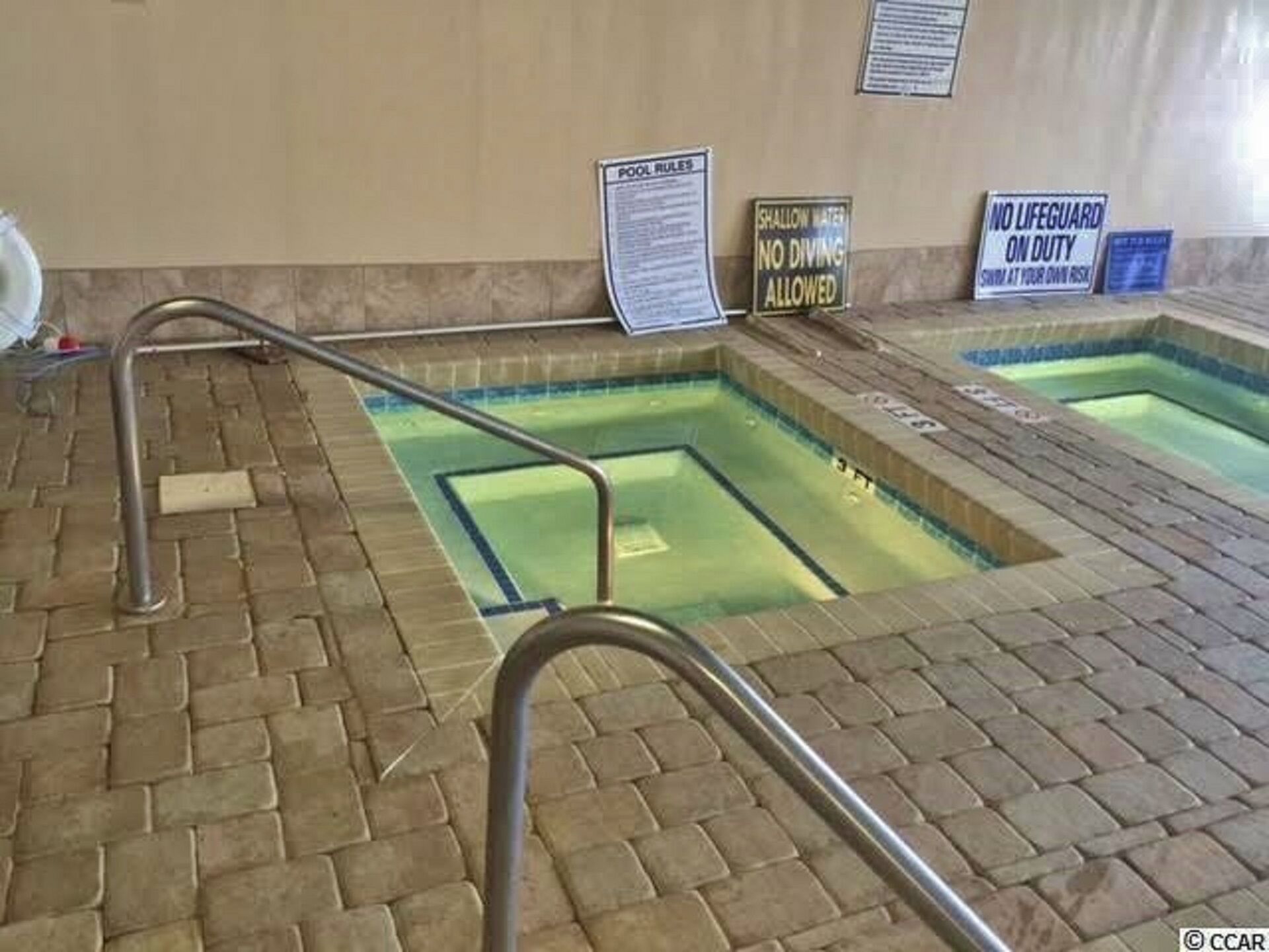 Un espace piscine doté de rampes métalliques, de panneaux d’avertissement indiquant la faible profondeur de l’eau et d’un panneau indiquant la présence d’un sauveteur.