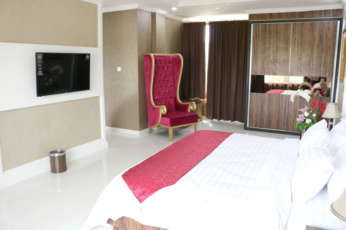A hotel room with a large bed, a red upholstered chair, a television mounted on the wall, and a sliding door closet.