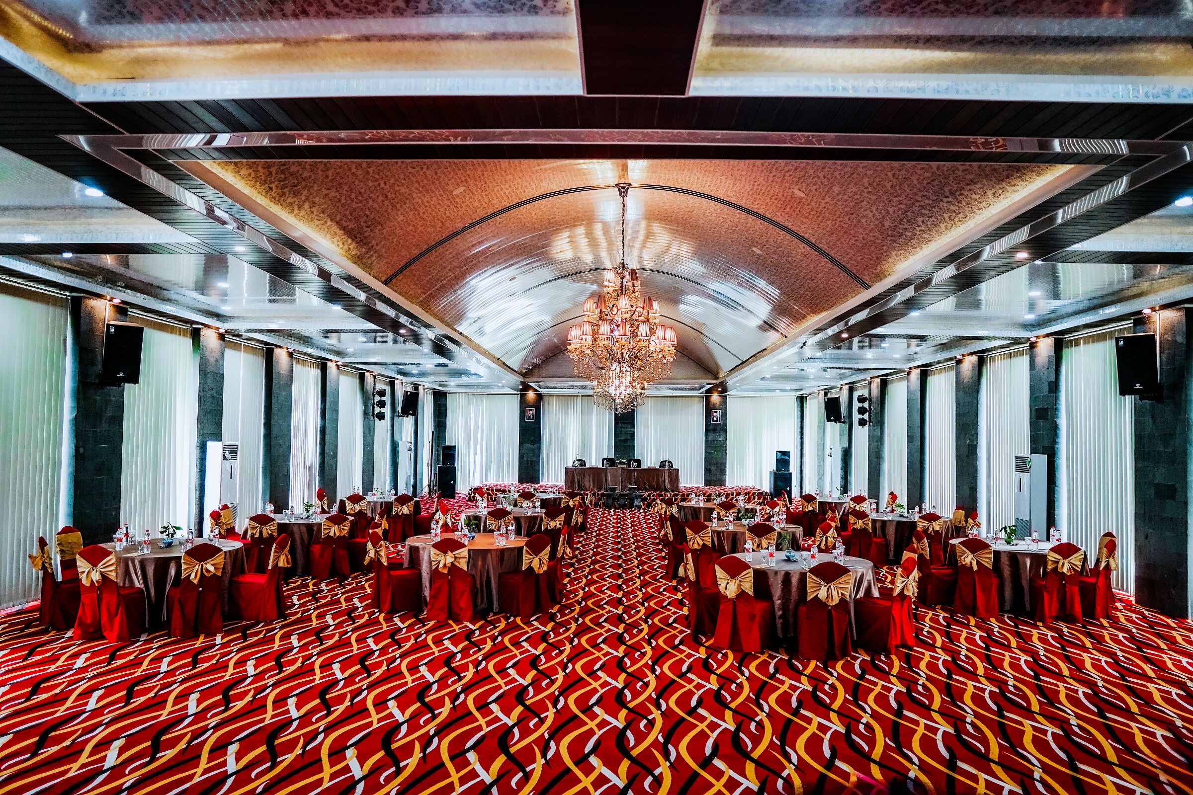 A banquet hall with rows of round tables, red carpet flooring, and a central chandelier.