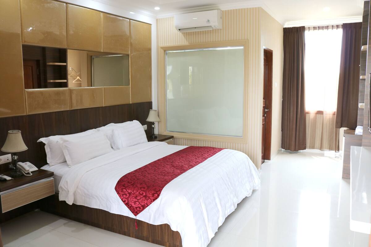A hotel room with a large bed, a bedside table with a lamp, a mirror, a wardrobe, and a large window with curtains.